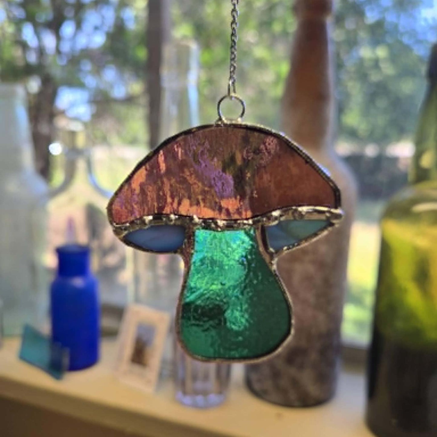 A stained glass mushroom suncatcher hanging from a chain, featuring a textured copper-brown cap, blue-green glass stems, and dark green glass base, set against a softly blurred background of bottles and greenery.