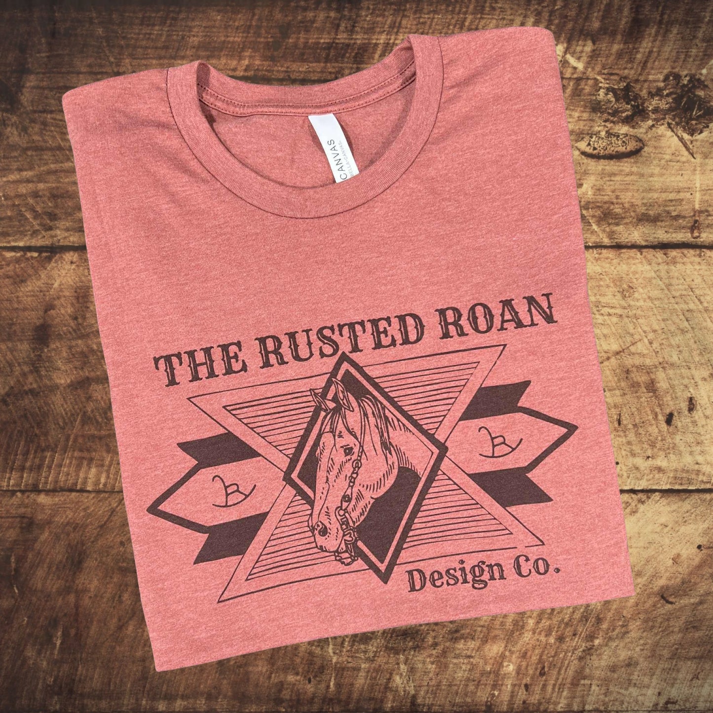 Folded heather clay t-shirt with 'The Rusted Roan Design Co.' logo on a wooden surface