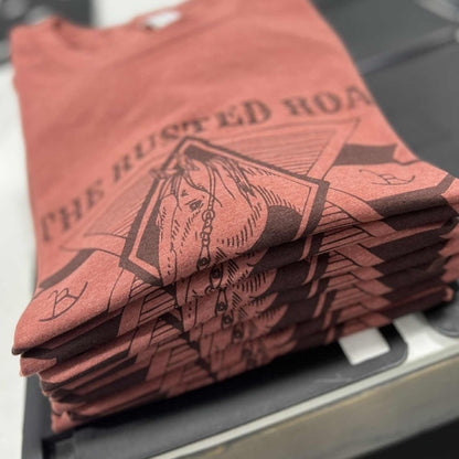 Pile of folder heather clay color tees with The Rusted Roan Logo
