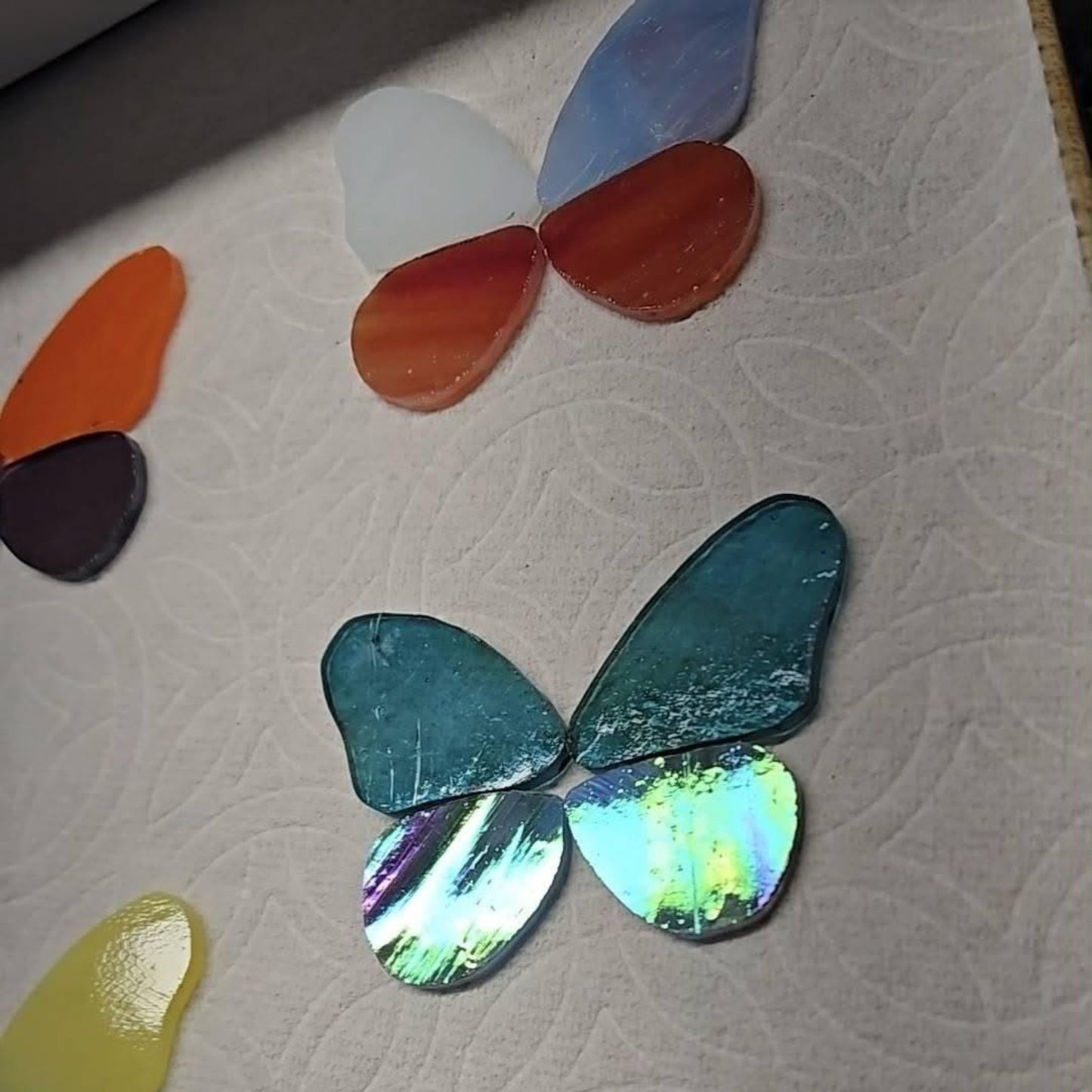 A close-up of butterfly wing-shaped pieces arranged on a textured white surface. The main visible piece is iridescent teal-green with blue-green and rainbow reflections, while other partial pieces in orange, white, blue, red, yellow, and dark brown are visible around it.