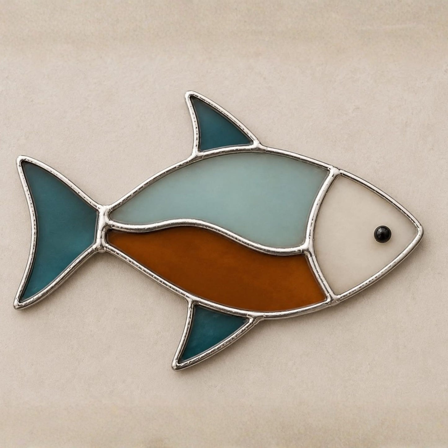 A stained glass fish ornament with a silver metal frame, featuring a light blue top, a brown lower section, a white face, and teal fins, displayed on a neutral beige background.